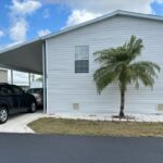 White mobile home with a palm tree and carport under a blue sky.